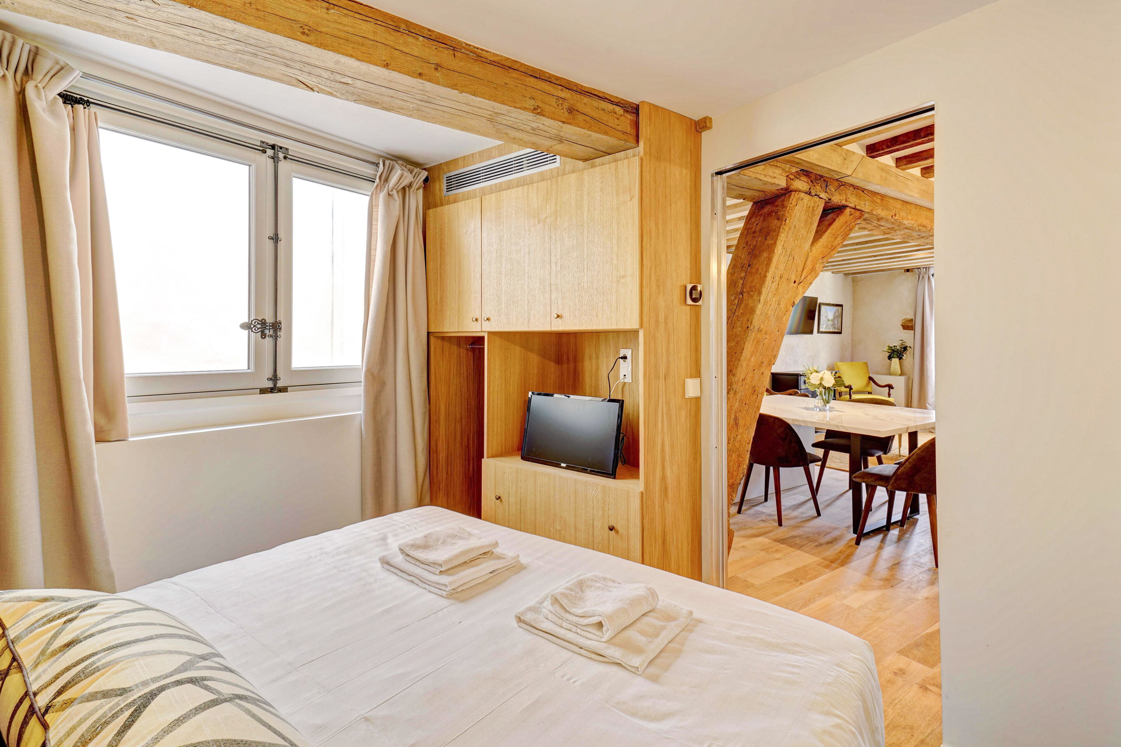 Quarto com cama, TV sobre um aparador de madeira e um espaço de refeições ao fundo.