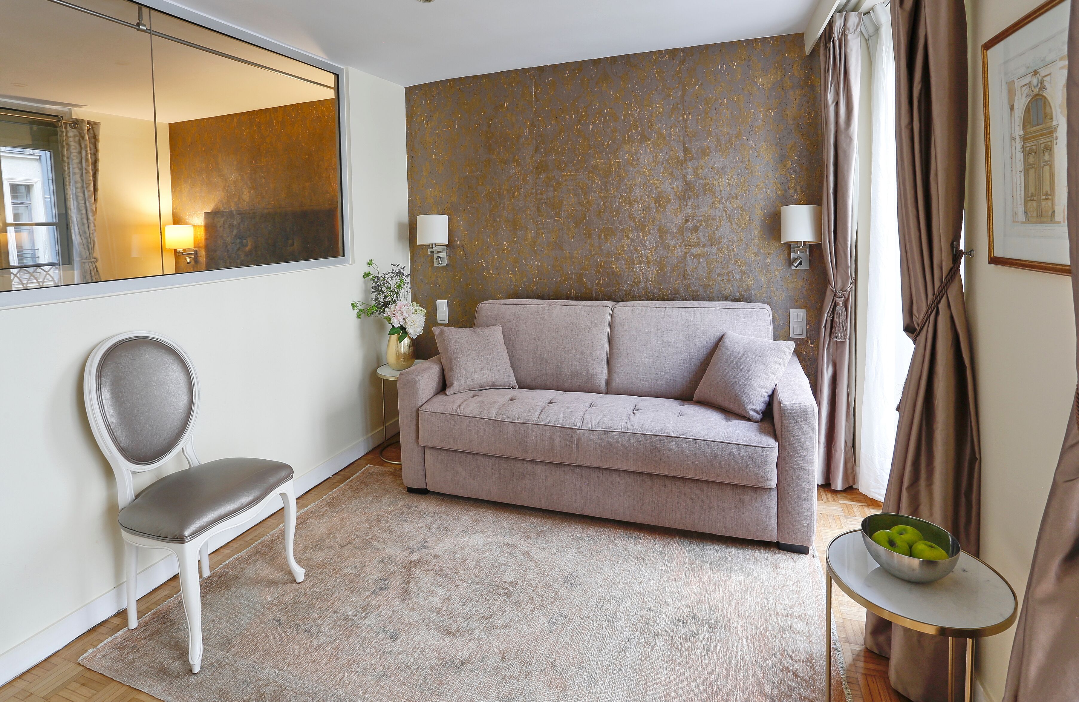 A living room with a sofa, a chair, a small table with a bowl of apples, and a wall-mounted lamp.