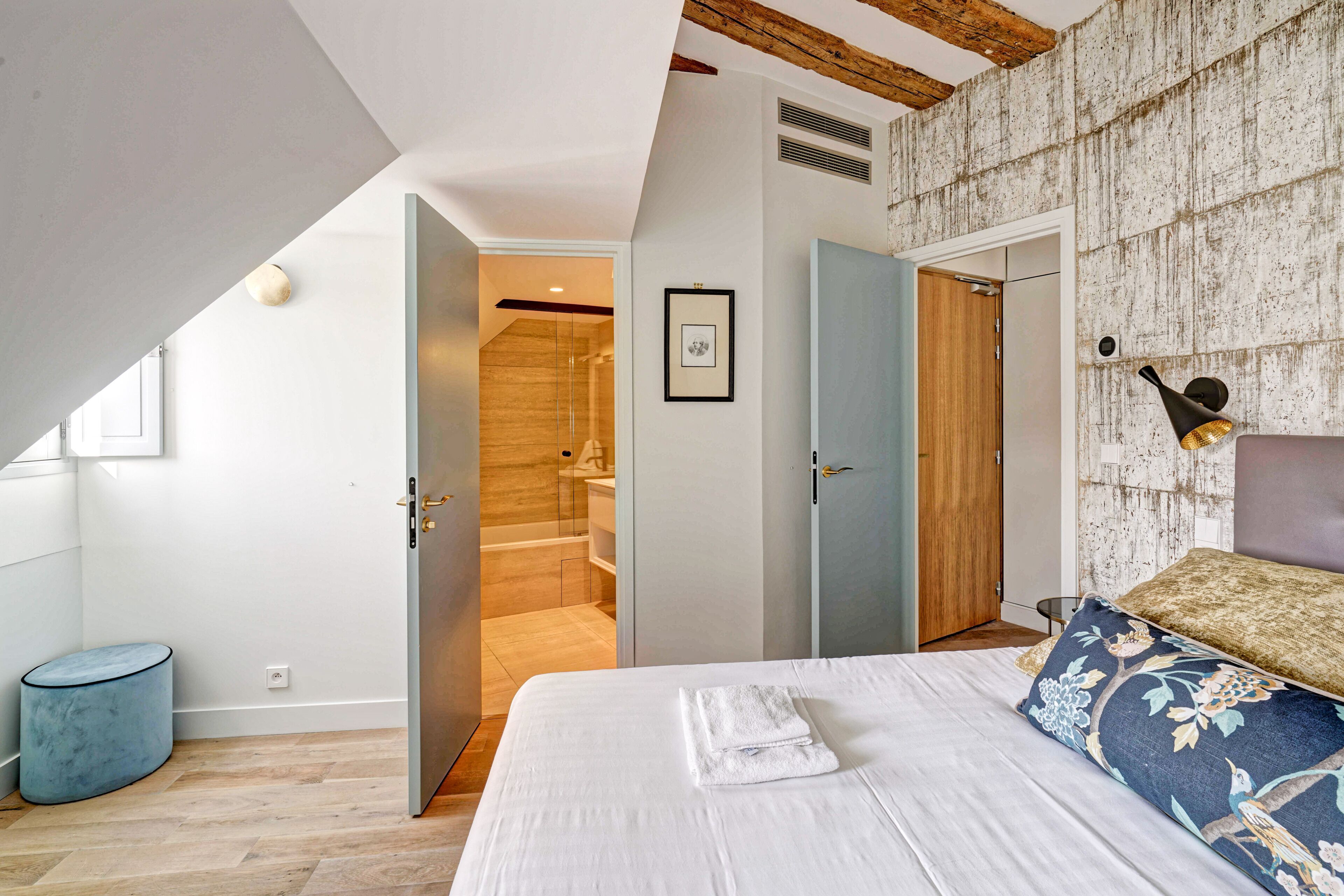 A bedroom with a bed, a blue ottoman, a wooden ceiling, and a bathroom visible through an open door.