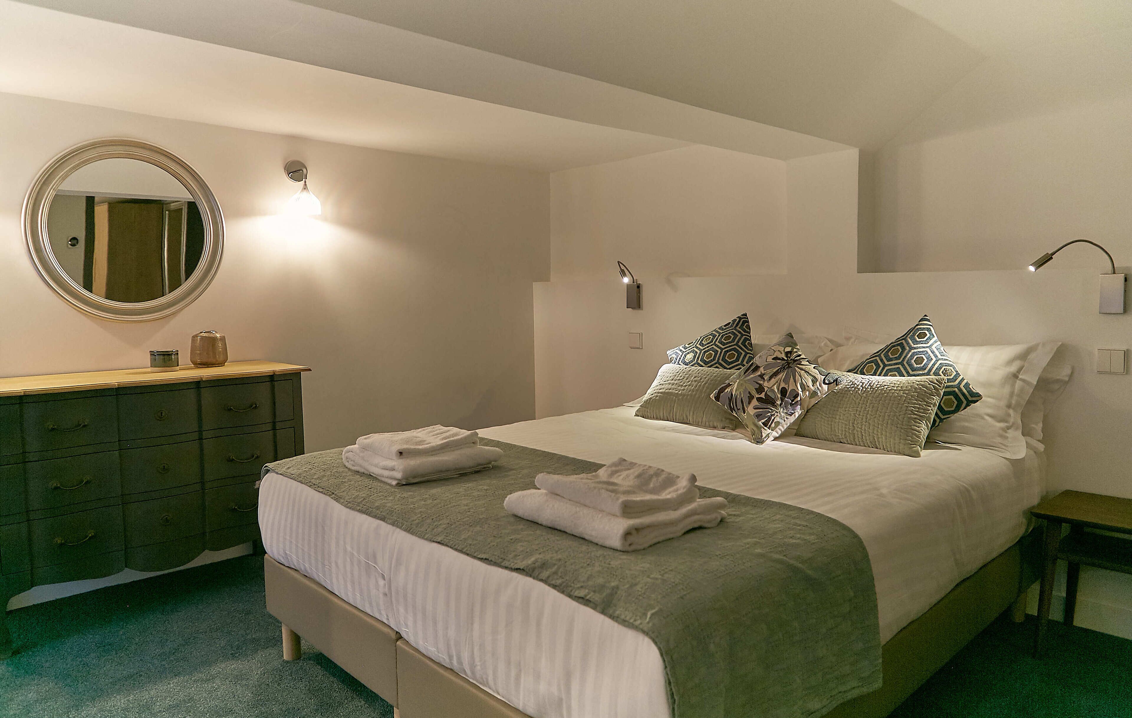 A neatly made bed with pillows and folded towels, a round mirror above a dresser, and wall-mounted lights.