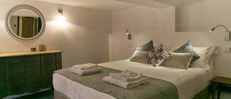 A neatly made bed with pillows and folded towels, a round mirror above a dresser, and wall-mounted lights.