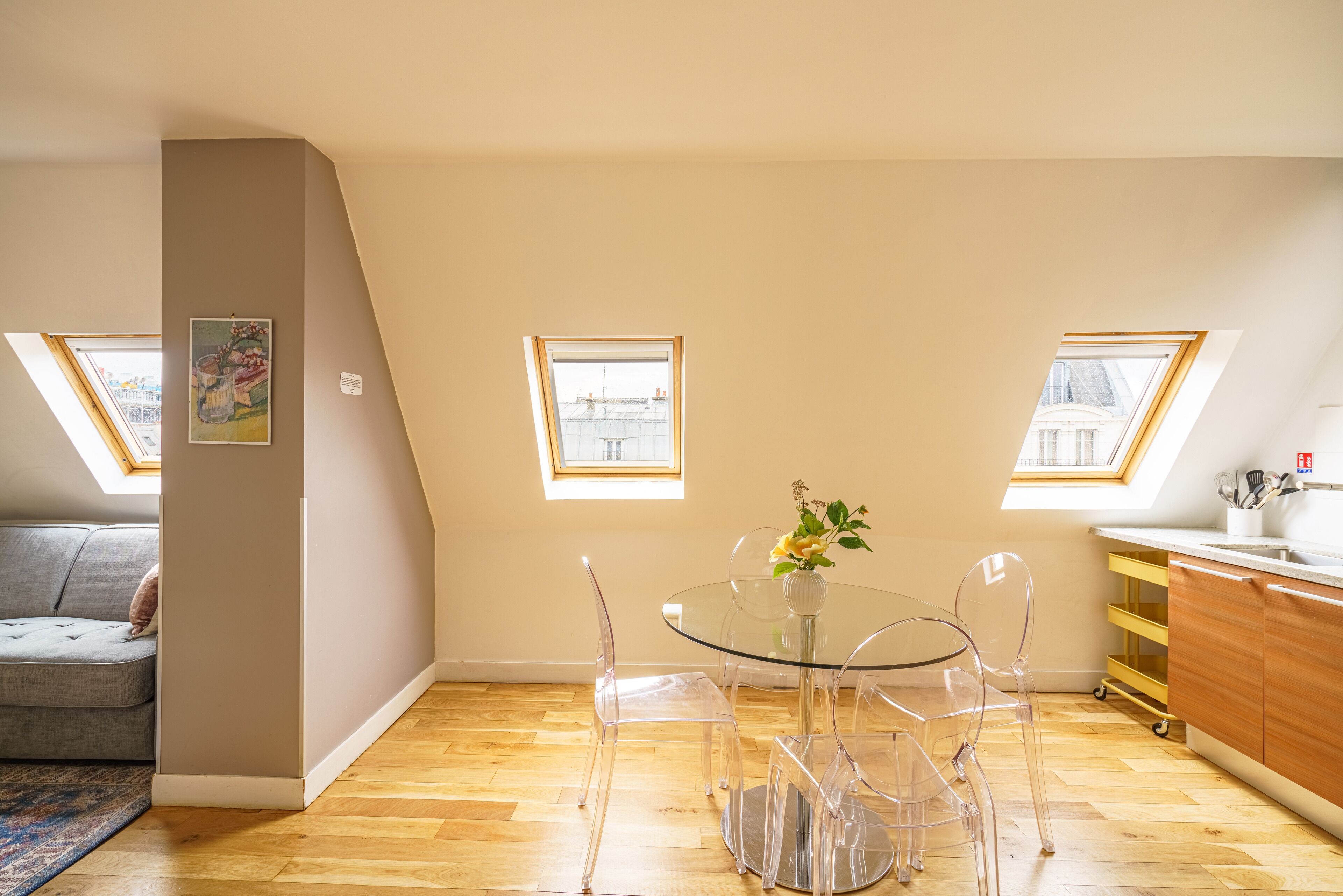 A modern loft apartment with a dining area featuring a glass table and chairs, a kitchen with wooden cabinets, and skylights providing natural light.