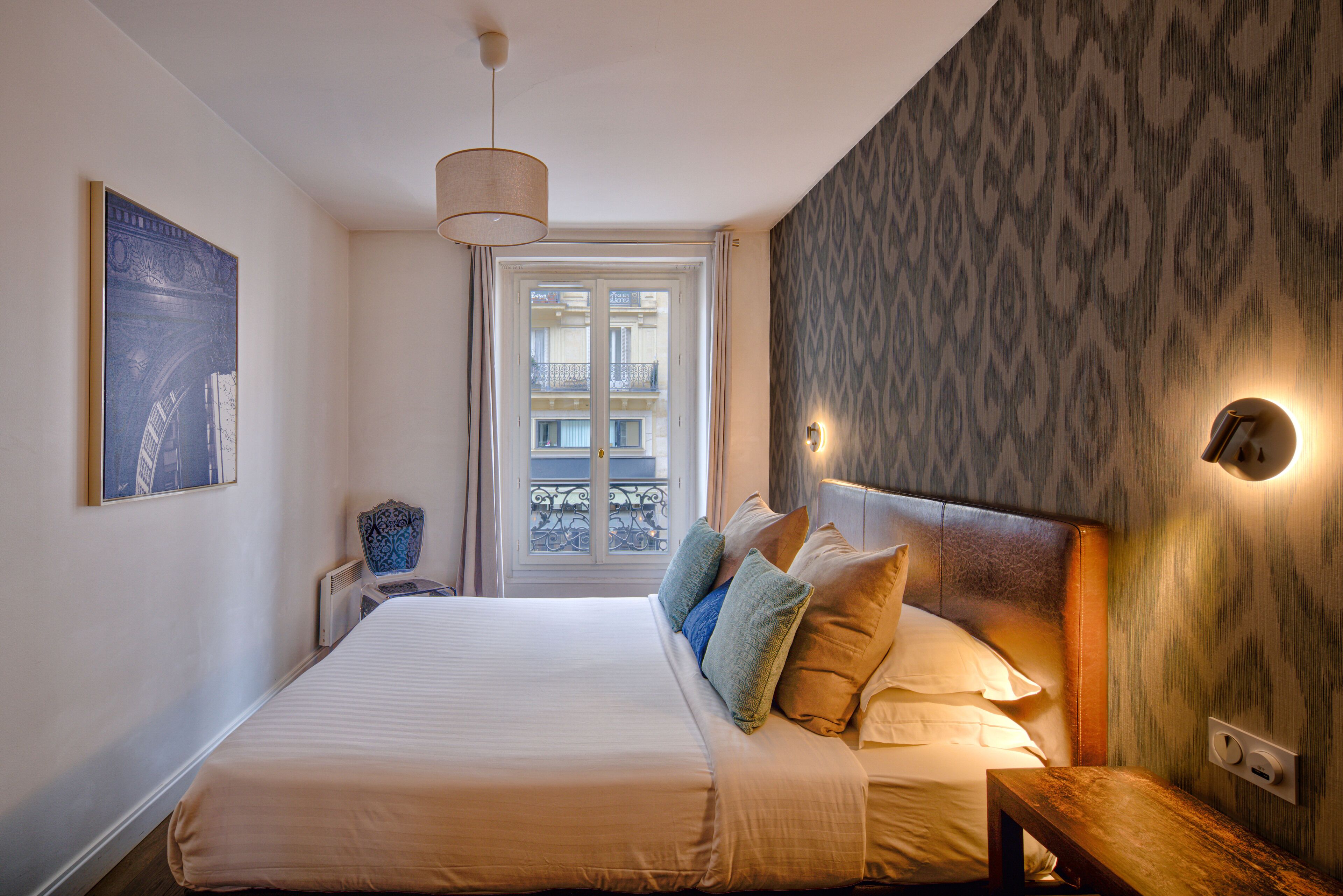 A neatly arranged bedroom with a large bed, a bedside table, a lamp, and a window with a view of a balcony.