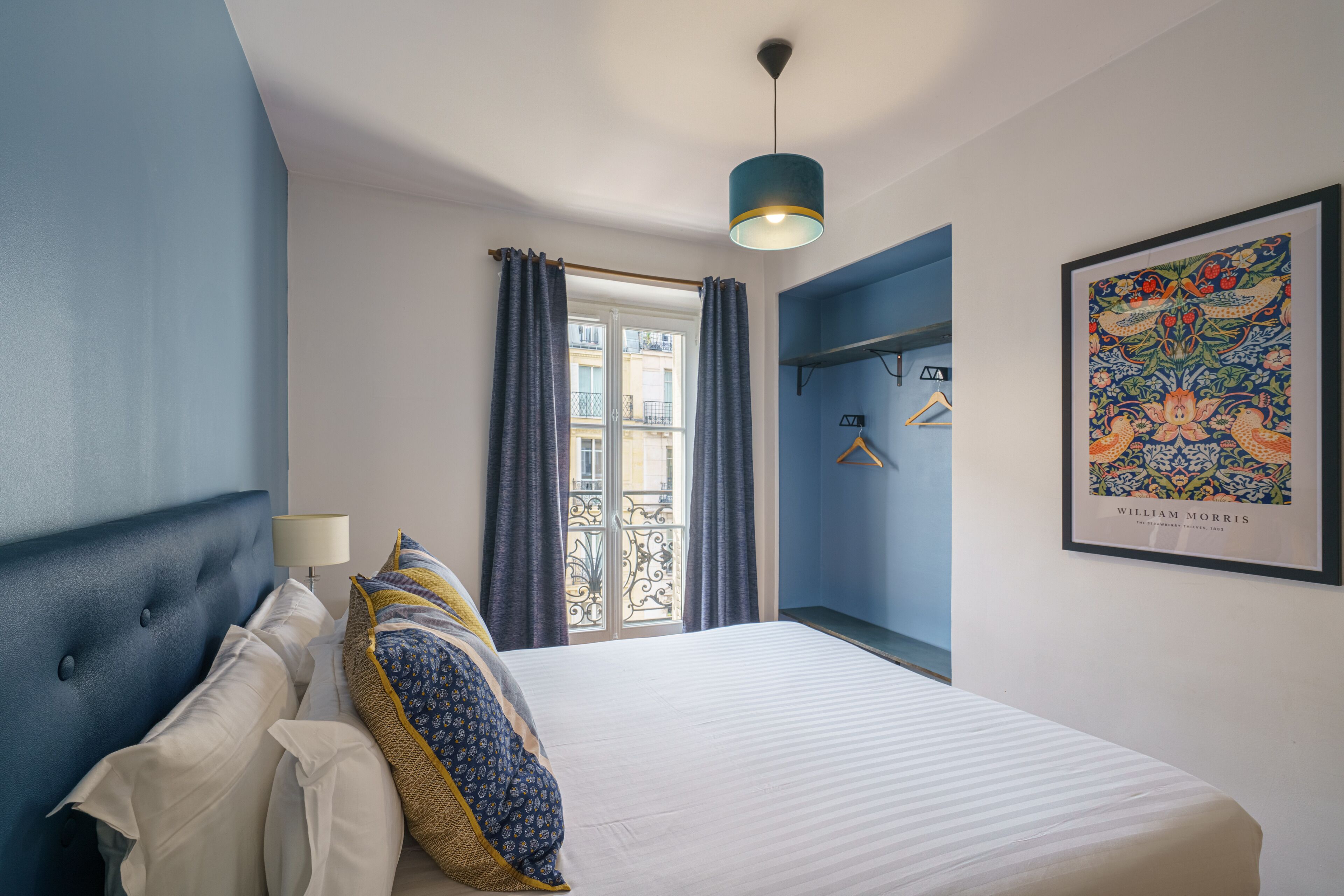 A bedroom with a large bed, a hanging light fixture, a window with curtains, and a framed picture on the wall.