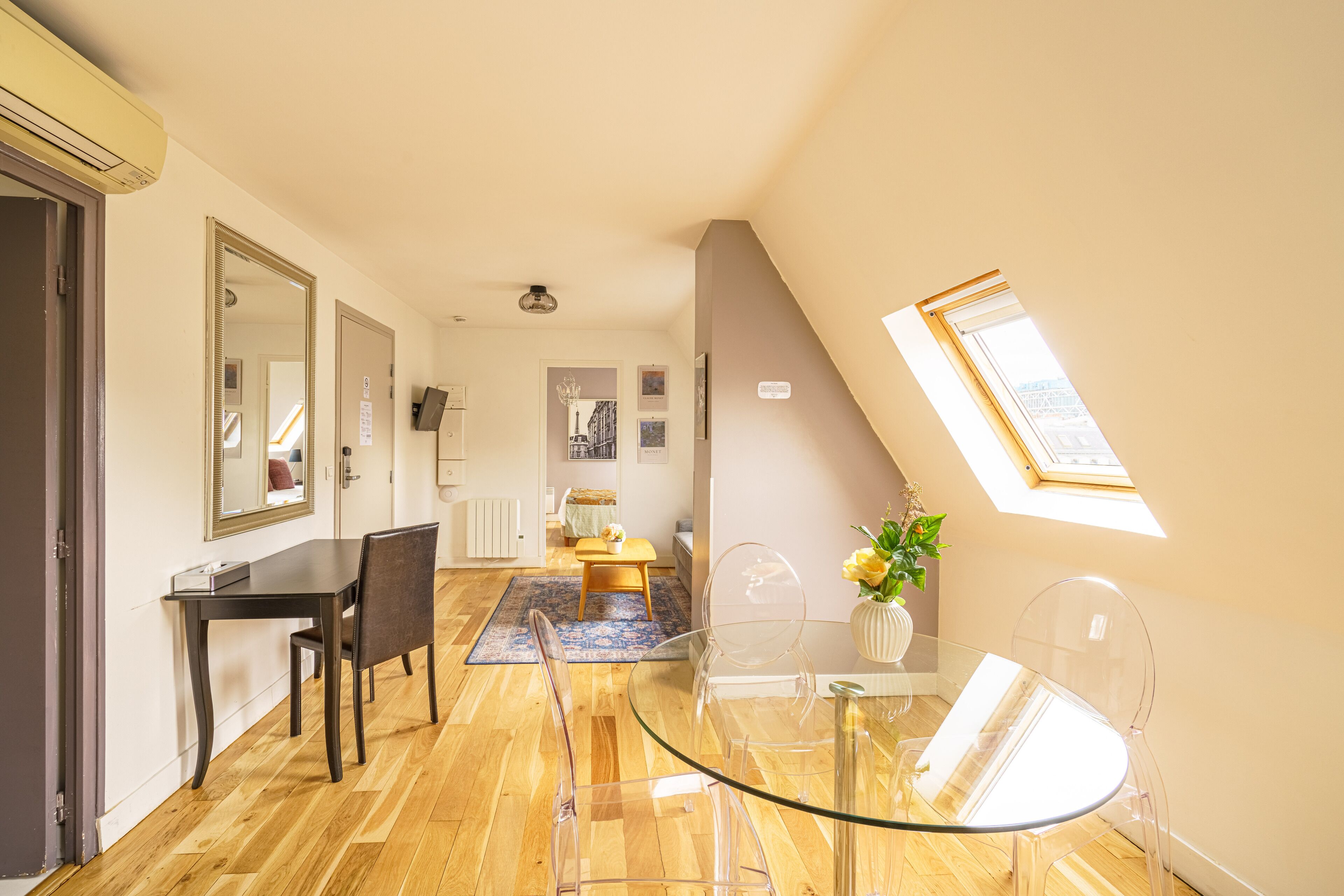 A modern living room with a glass dining table, clear chairs, a wooden floor, a large mirror, a small sofa, and a skylight.