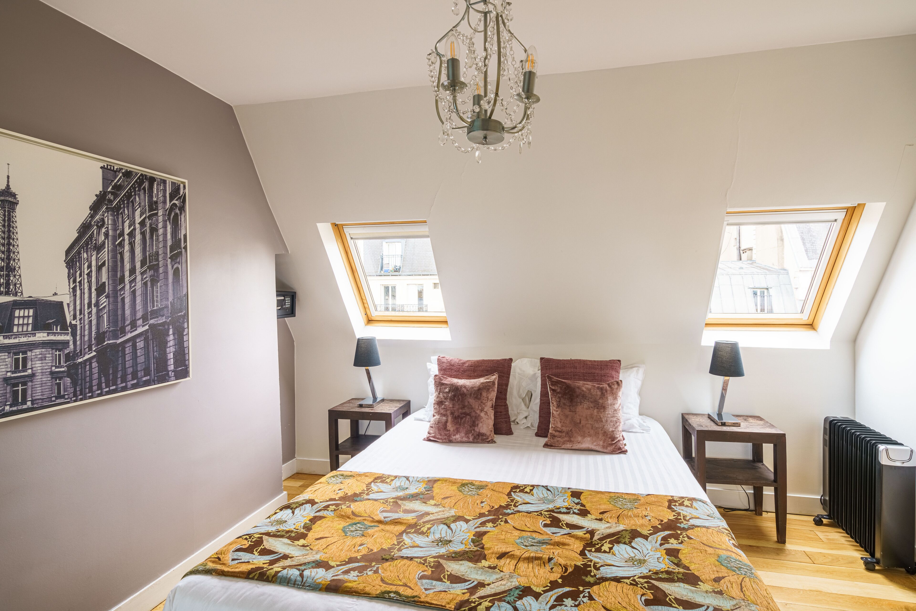 A bedroom with a large bed, two bedside tables, a chandelier, and a cityscape painting on the wall.