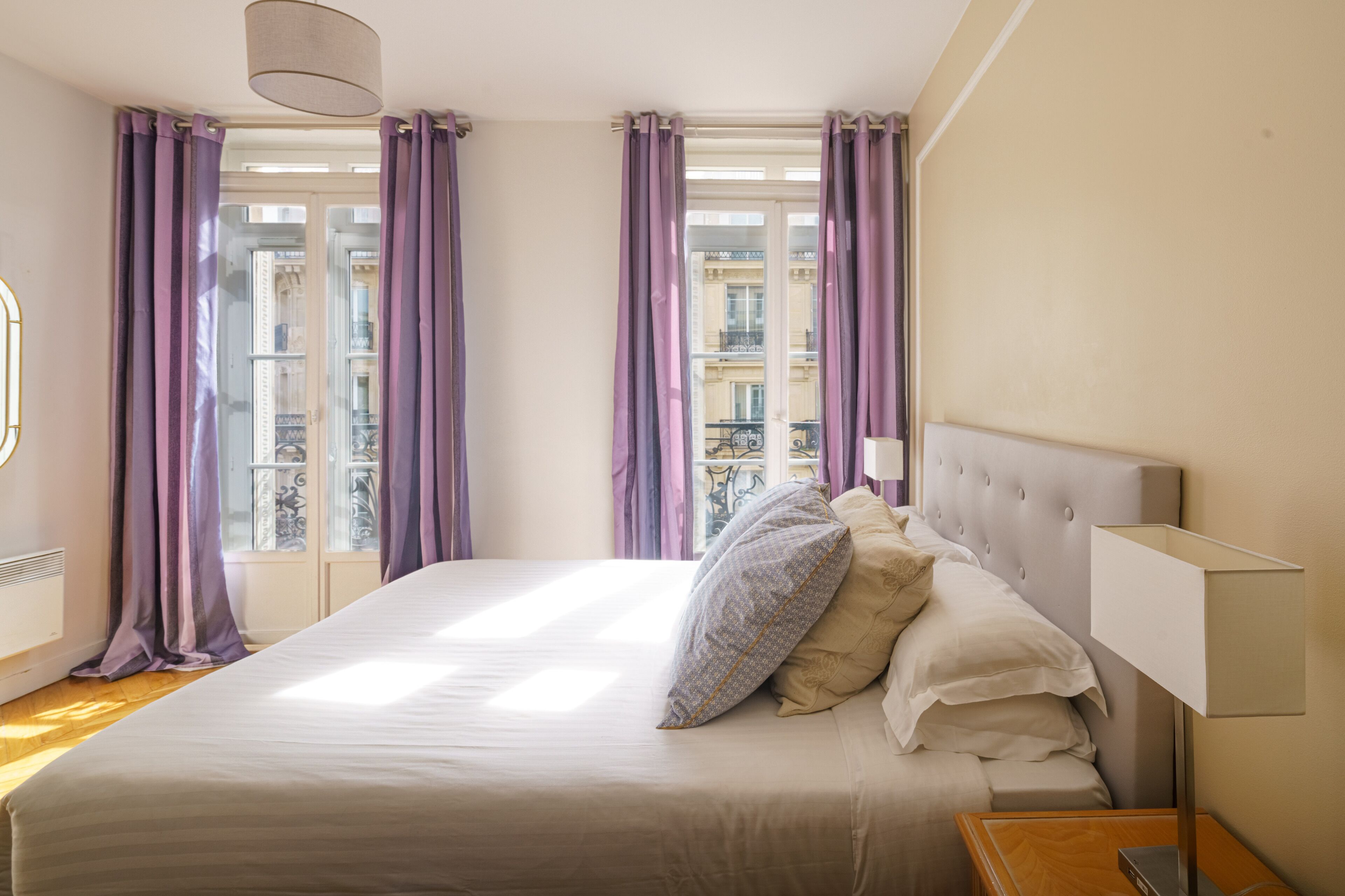 A hotel room with a large bed, a nightstand, a lamp, and a view of buildings through the window.
