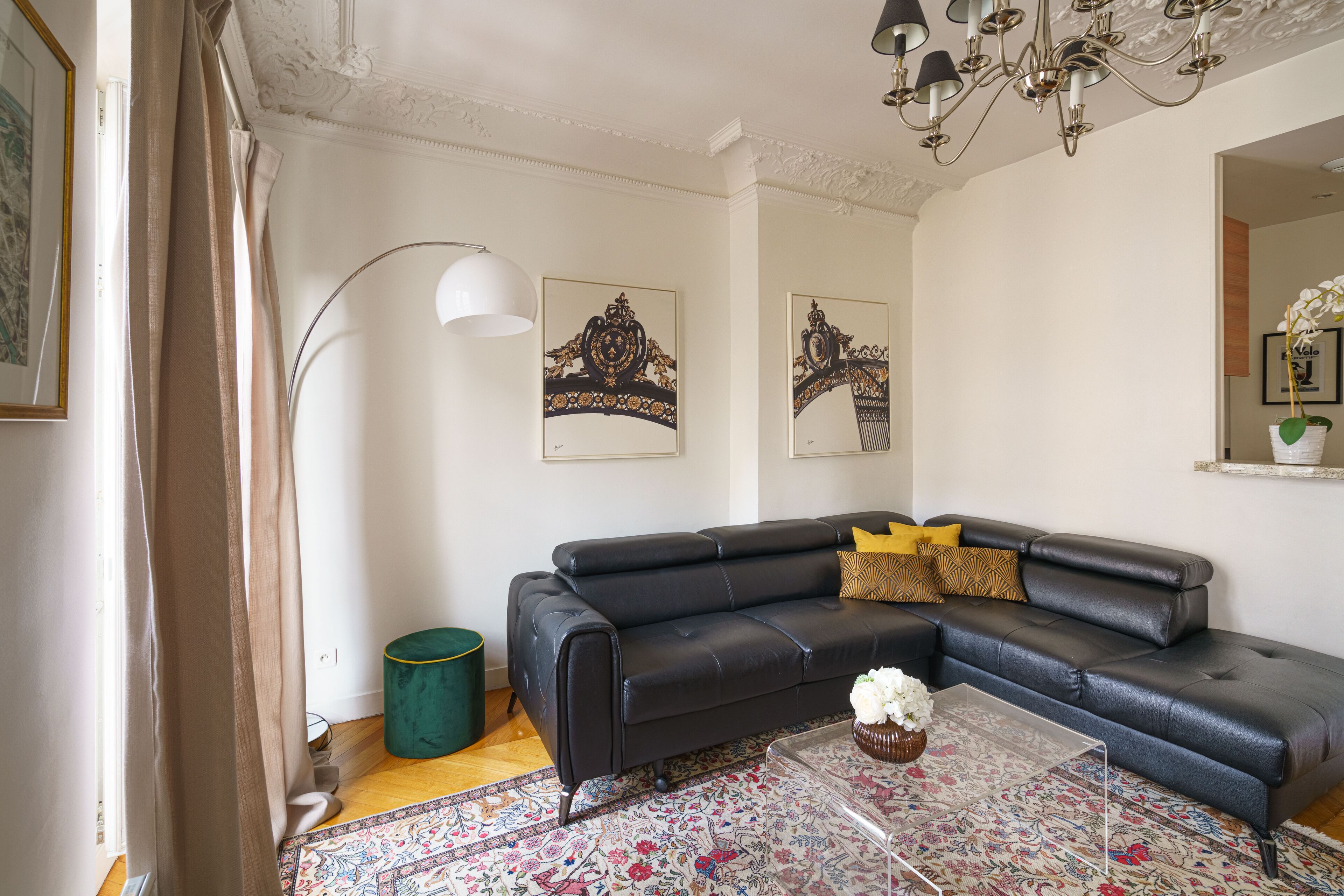 Een woonkamer met een zwarte leren bank, een transparante salontafel, een getextureerd vloerkleed en een groene poef.