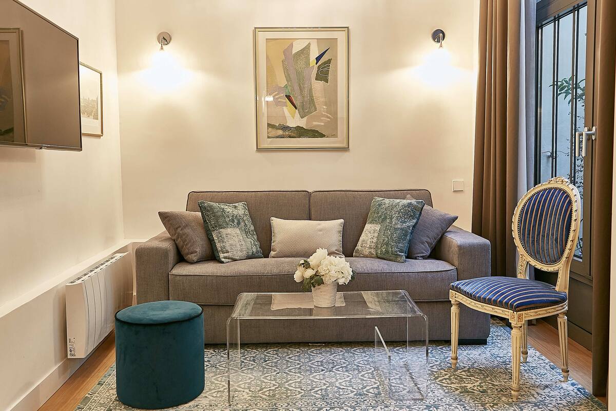 A living room with a grey sofa, a clear coffee table, a blue ottoman, a decorative chair, and a framed artwork on the wall.