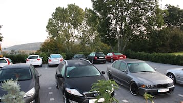 En parkeringsplads med flere biler, heriblandt en sort sedan og en sølvfarvet sedan, omgivet af træer og et kuperet landskab i baggrunden.