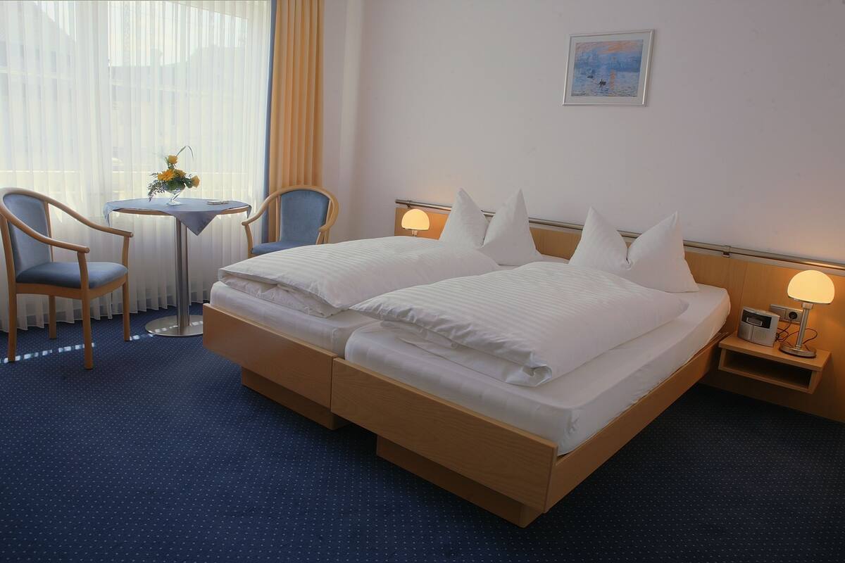 Une chambre d’hôtel avec un lit double, deux tables de chevet, une petite table avec un vase, deux chaises et une fenêtre avec des rideaux.