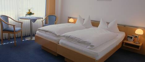Habitación de hotel con cama doble, dos mesitas de noche, una mesita con un florero, dos sillas y una ventana con cortinas.
