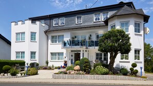 A two-story hotel with a balcony on the second floor, a well-maintained garden, and a clear blue sky. - Hotel Retterath am Nürburgring (Wiesemscheid)