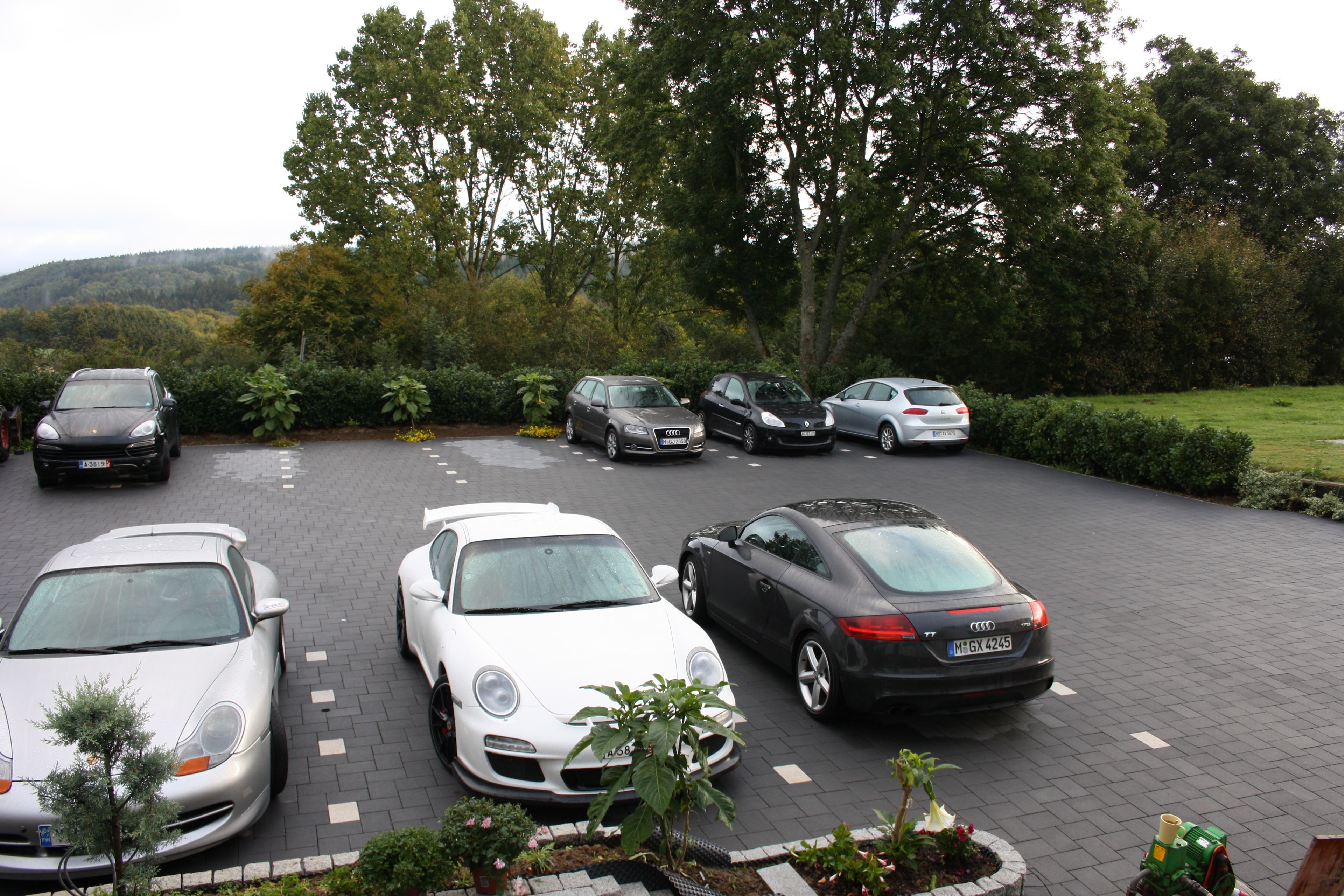 A parking lot with several cars, including a white Porsche and a black Audi, surrounded by greenery and trees.