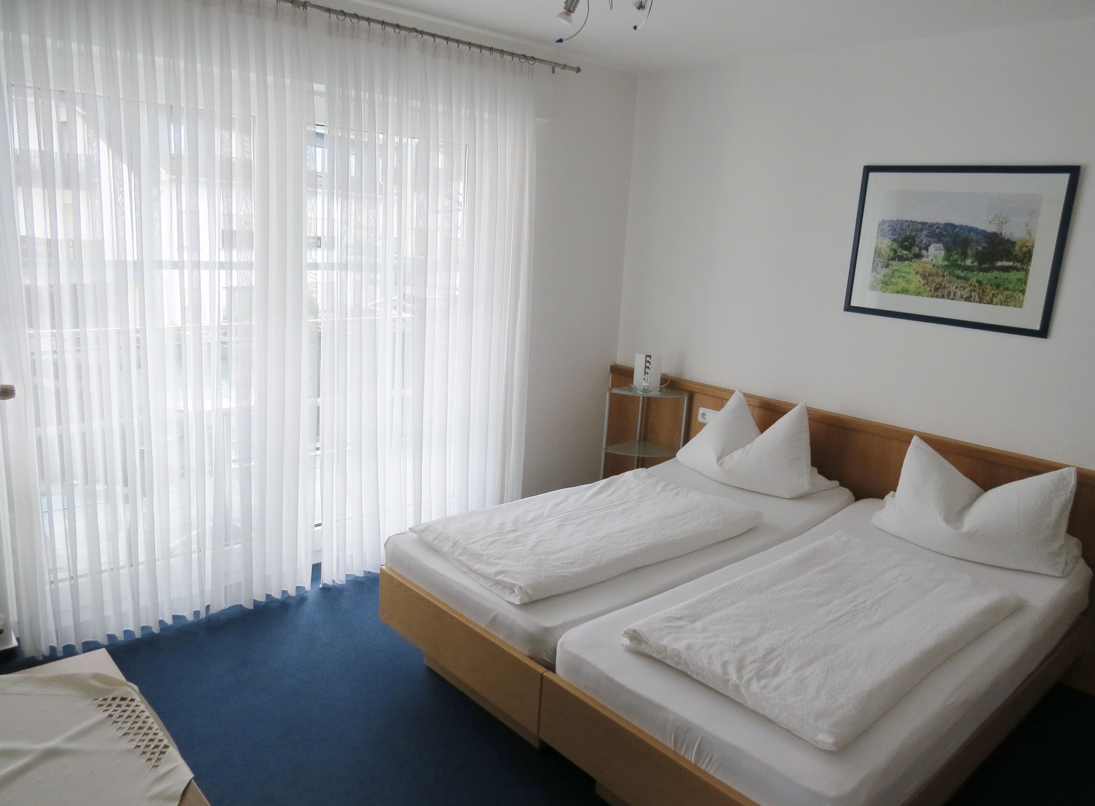 Habitació d'hotel amb dos llits, una catifes blava, cortines blanques, una tauleta de nit amb una làmpada i un quadre al mur.
