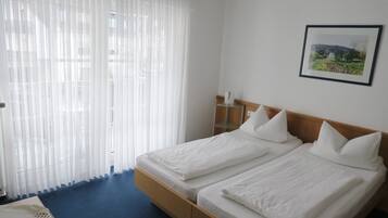 Habitació d'hotel amb dos llits, una catifes blava, cortines blanques, una tauleta de nit amb una làmpada i un quadre al mur.