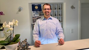 A man standing behind a reception desk in a hotel lobby with a framed award sign, a potted orchid, and a decorative metal sculpture. - Hotel Retterath am Nürburgring (Wiesemscheid)