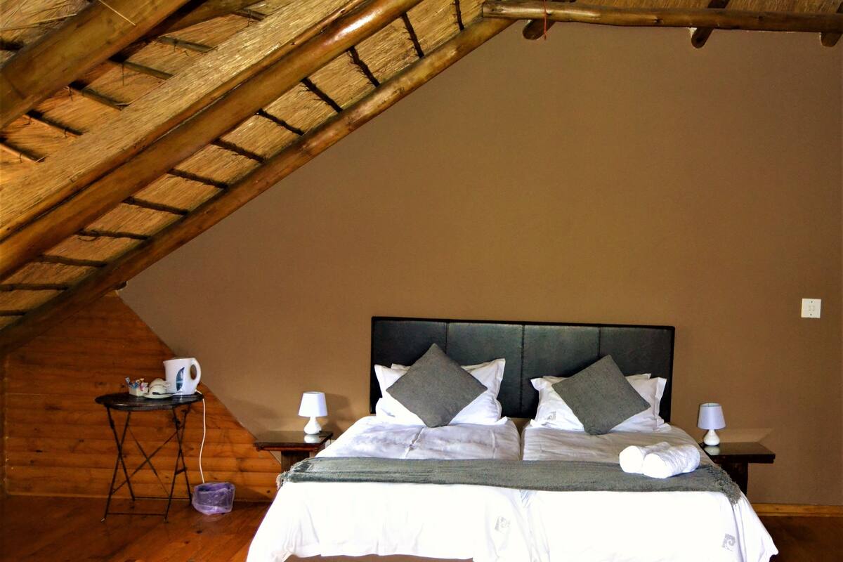 A bedroom with a sloped ceiling, a bed with pillows, a nightstand with a lamp, and a small table with a cup and a lamp.