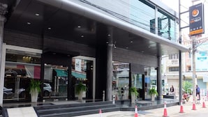 A modern hotel entrance with a covered walkway, potted plants, and a visible car in the background. - The Grand Day Night (Pattaya)