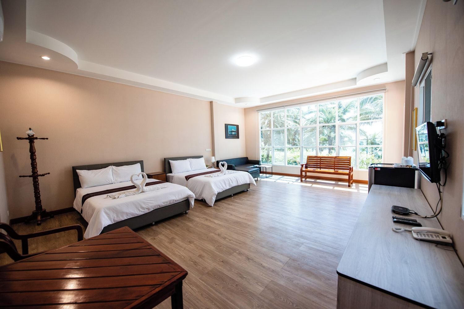 A hotel room with two beds, a desk, a chair, a wooden table, a TV, and a large window with a view of greenery.