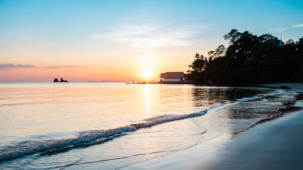 Une scène de plage avec un coucher de soleil, une eau calme et un bâtiment sur le rivage.