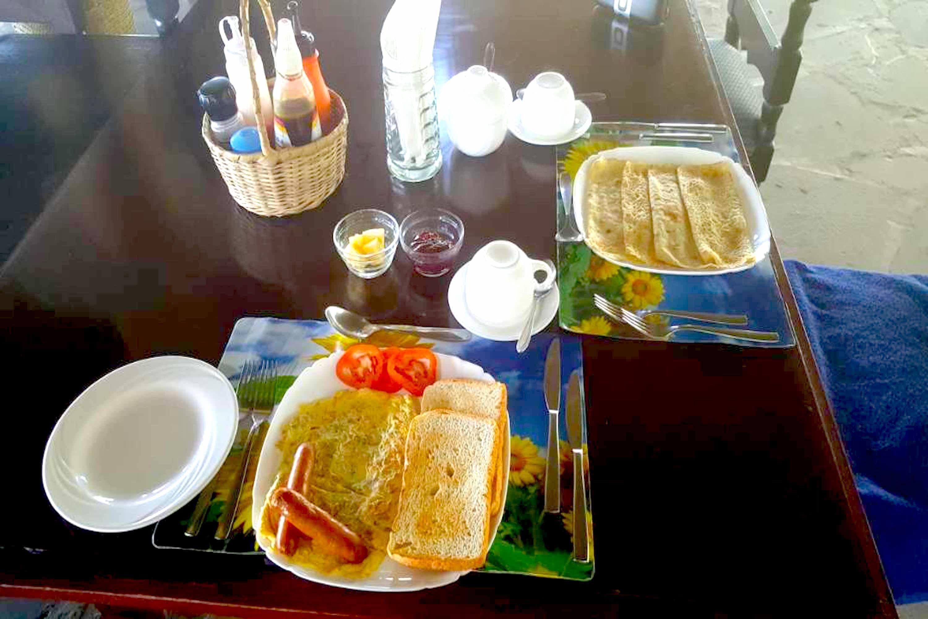Een ontbijttafel met een bord eieren, toast en tomaten, een mand met toevoegingen, en een theepot.