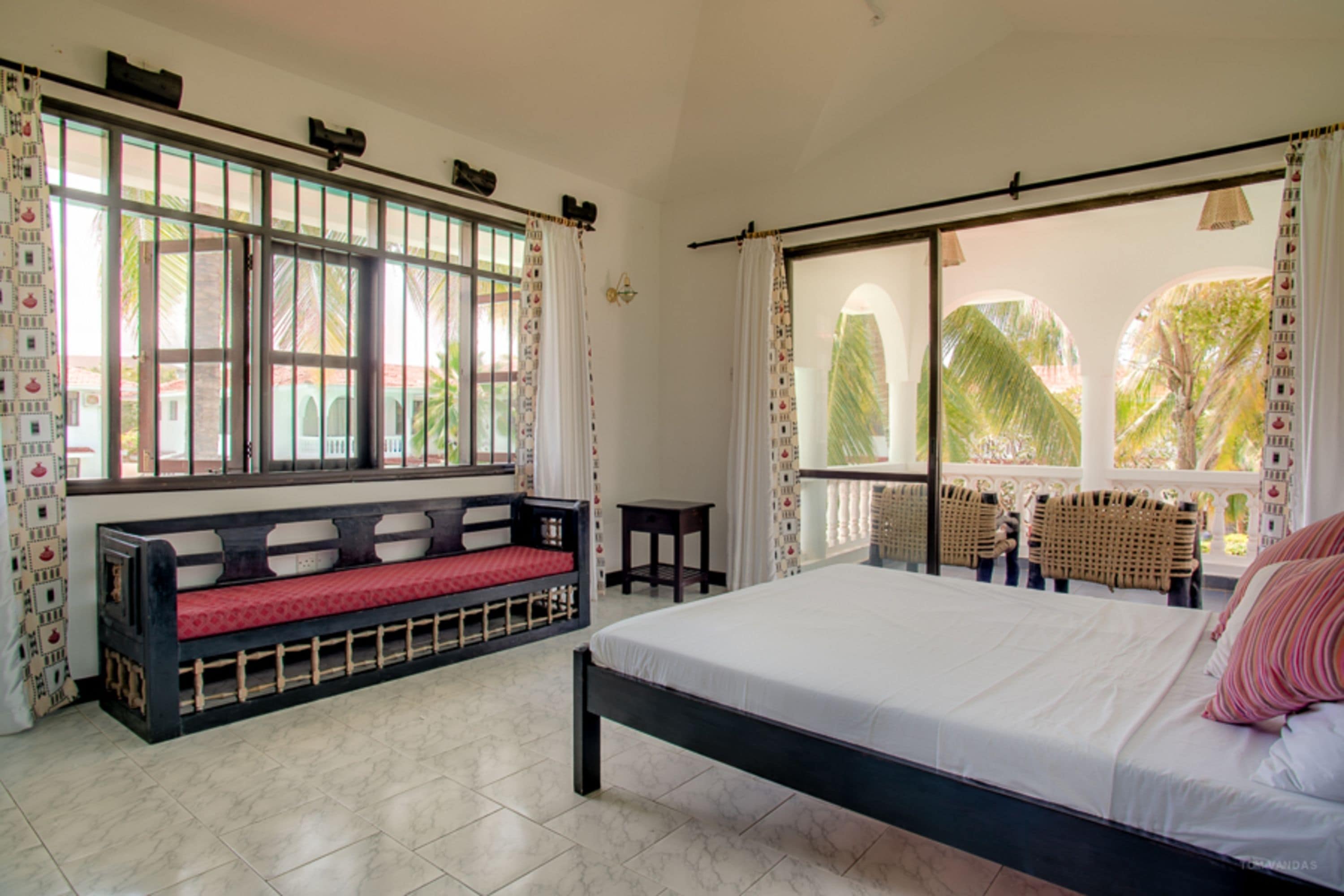 Un dormitorio con una cama grande, un banco con un cojín rojo y vistas a palmeras a través de la ventana.