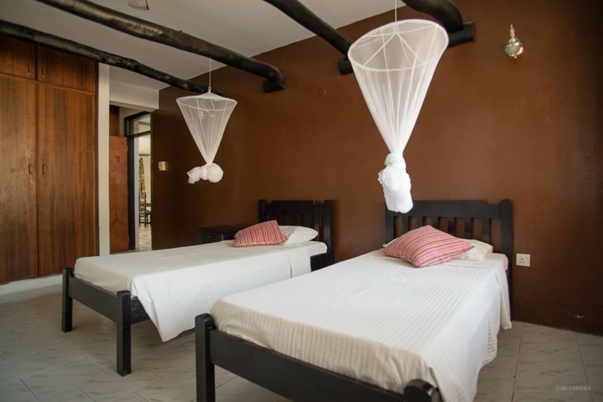 Dos camas individuales con sábanas blancas y almohadas a rayas, un armario de madera y una mosquitera colgante.