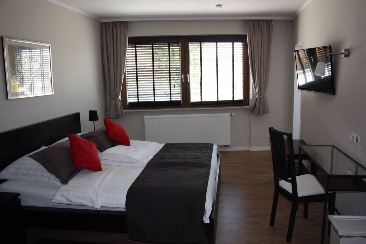 A bedroom with a bed, a desk, a chair, a TV, and a window with curtains.