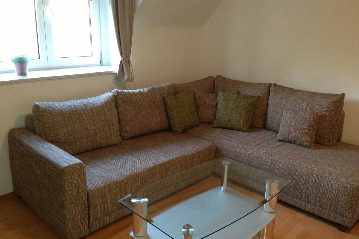 A brown sectional sofa with multiple cushions, a glass coffee table, and a window with curtains.
