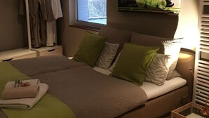 A bedroom with a bed, a window with blinds, a shelf with flowers, and a clothing rack. - WohnRaum Hotel & Gästehaus (Gusterath)