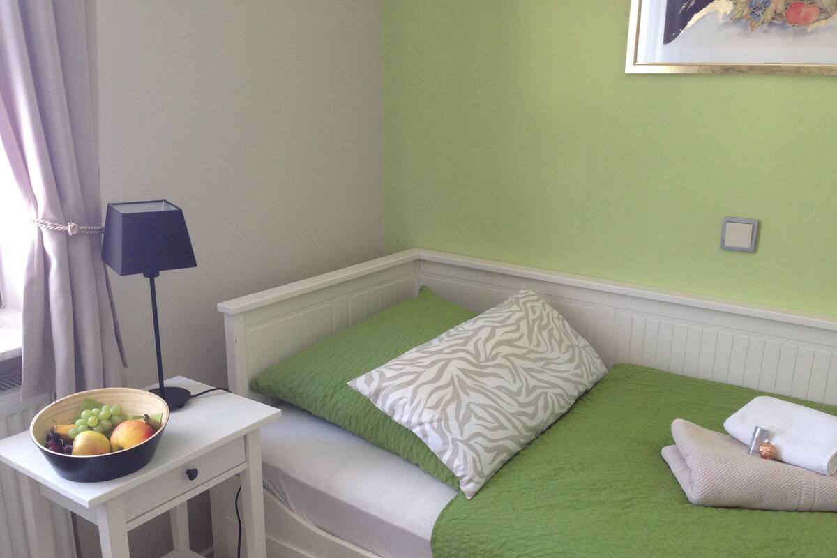 A small, single-bed room with a green bedspread and a zebra-patterned pillow. A white bedside table holds a bowl of fruit.