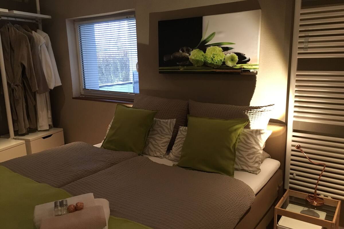 A bedroom with a bed, pillows, a nightstand, a lamp, a window, and a shelf with flowers.