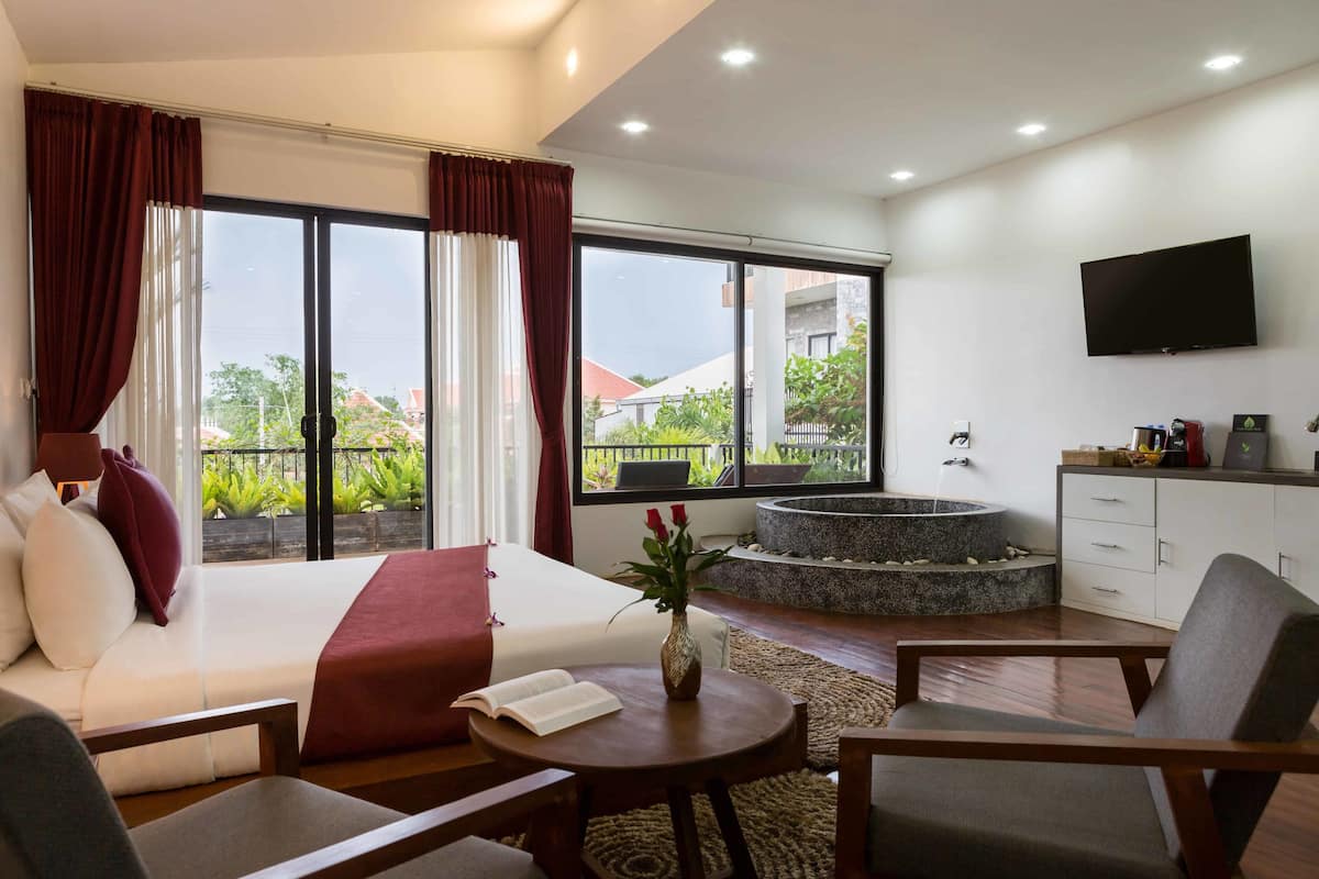 A modern hotel room with a large bed, a sitting area with a coffee table, a TV mounted on the wall, and a view of the outdoors through large windows.