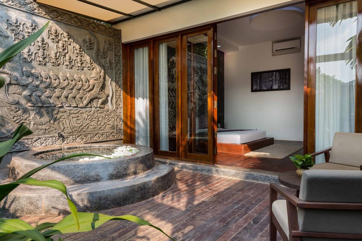 A modern interior with a stone relief wall, a stone fountain, a wooden deck, and a seating area with a sofa and armchairs.
