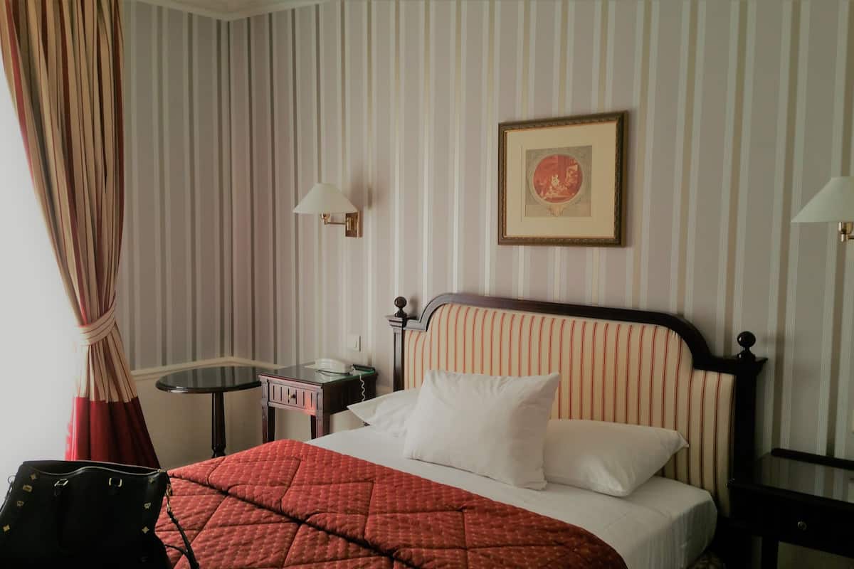 Ein Hotelzimmer mit gestreifter Tapete, einem Bett mit gestreifter Kopfteil, zwei Nachttischlampen, einem gerahmten Bild an der Wand, einem Telefon auf dem Nachttisch und einer roten, gesteppten Tasche auf dem Bett.