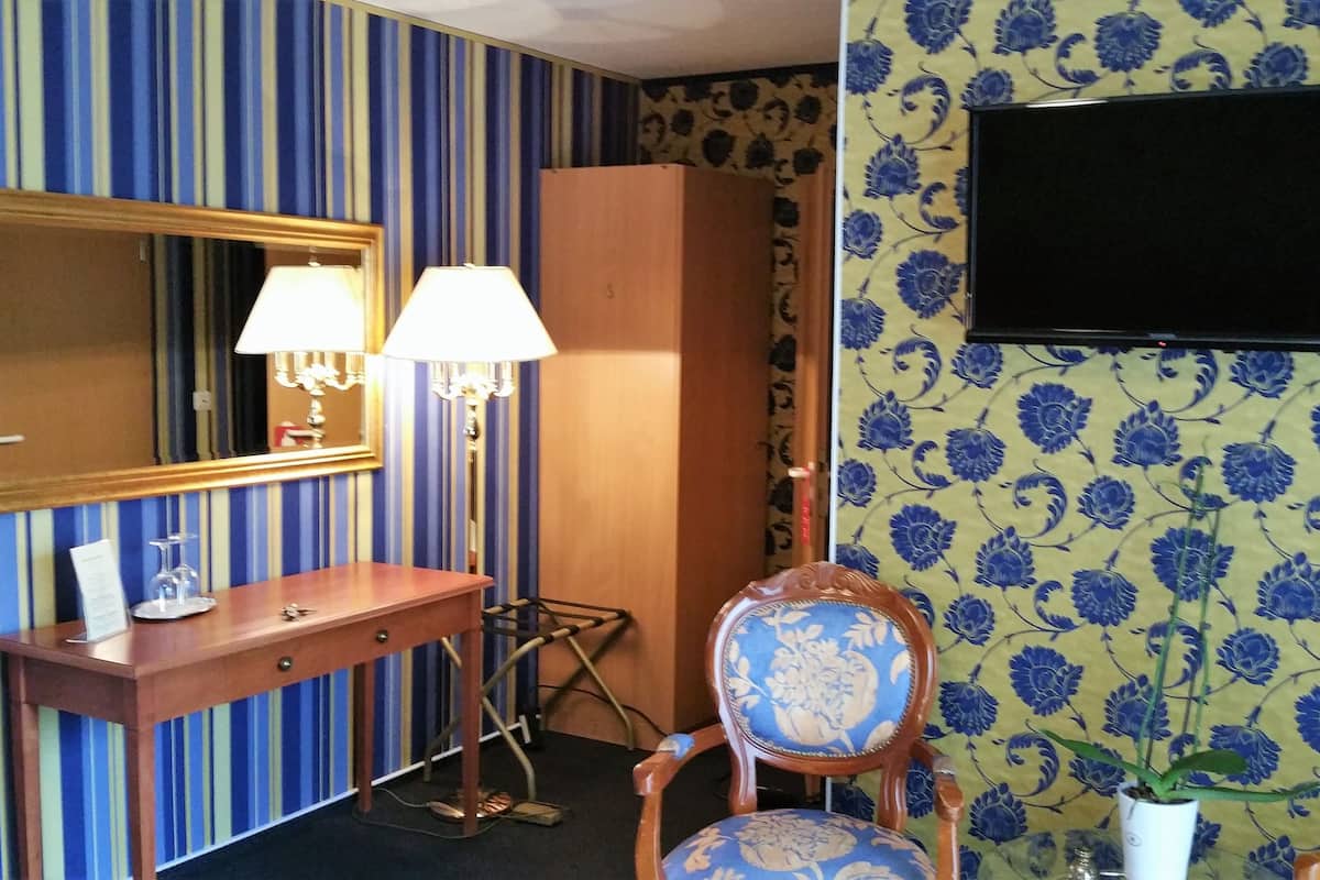 Ein Zimmer mit einem Holzanrichte, einem Spiegel, zwei Wandlampen, einem Fernseher, Tapete mit Muster und einem Sessel mit Blumenkissen.