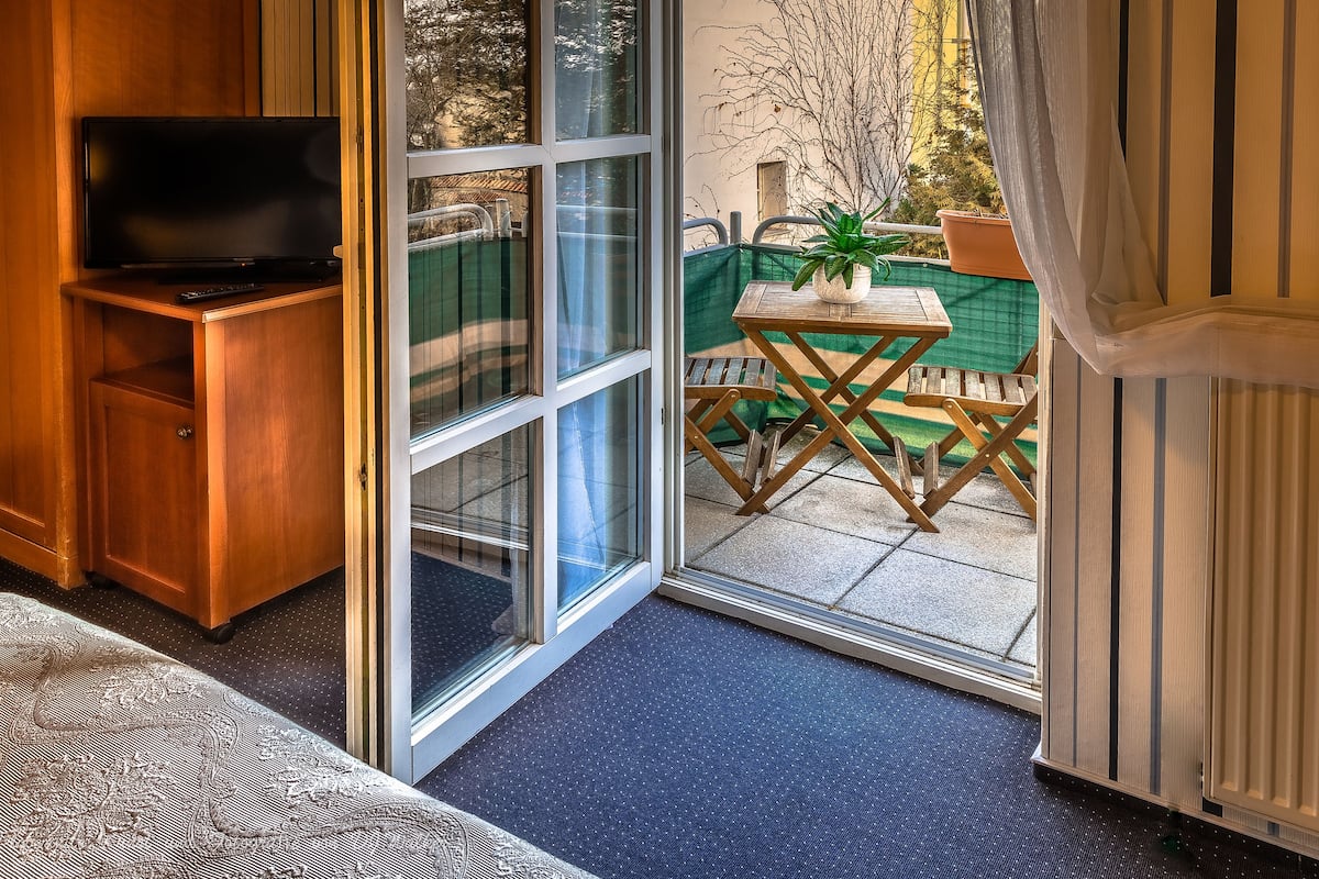 Ein Zimmer mit Fernseher auf einer Holzkassette, einer Schiebetür zu einem Außenbereich mit Tisch und Stühlen sowie einem Heizkörper an der Wand.