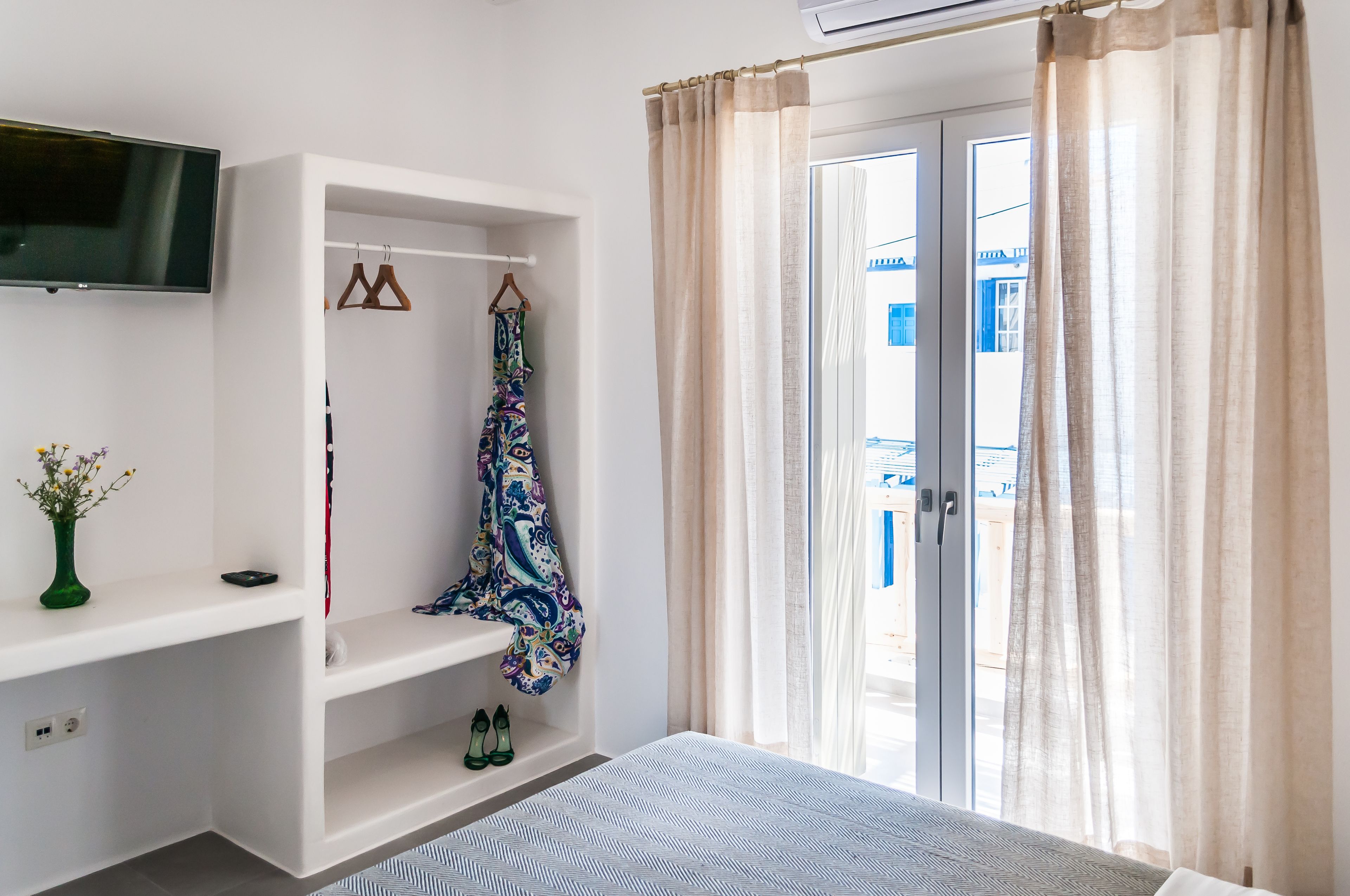 A hotel room with a bed, a TV mounted on the wall, a wardrobe, a shelf with shoes, and a window with curtains.