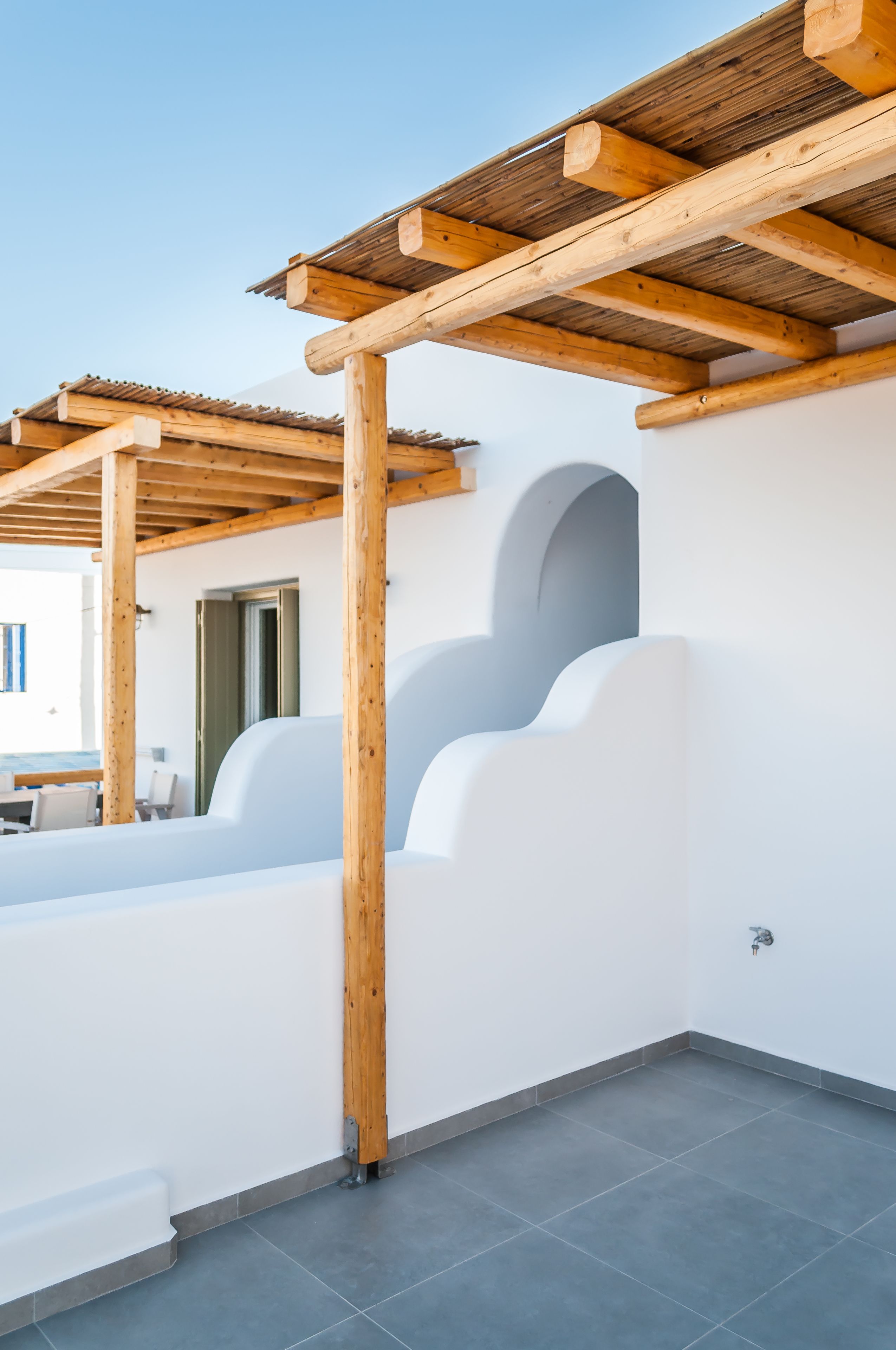 Una terraza moderna al aire libre con una pérgola de madera, paredes blancas y suelo de baldosas.