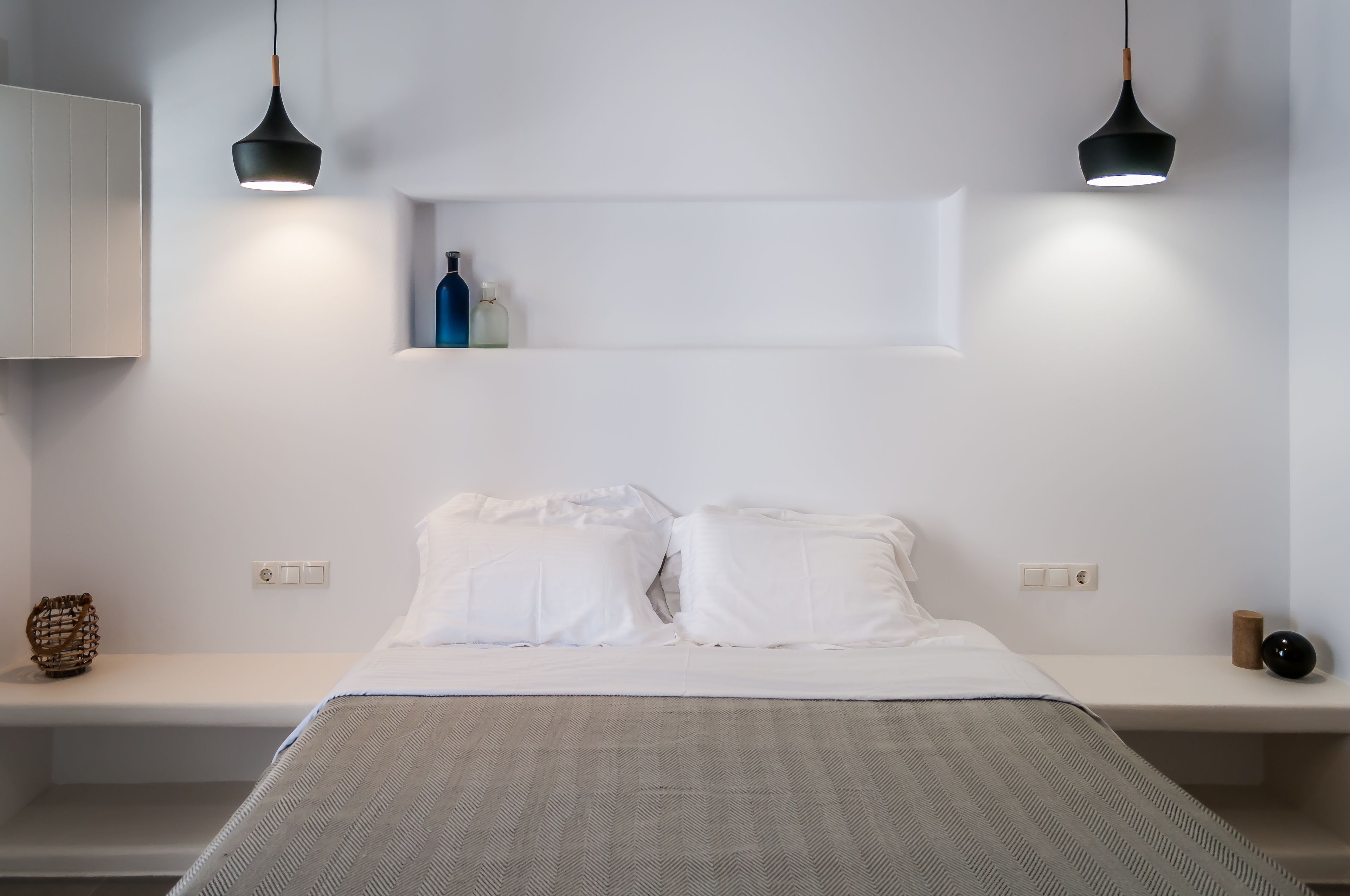 Un dormitorio minimalista con una cama, dos lámparas colgantes y una estantería con botellas.