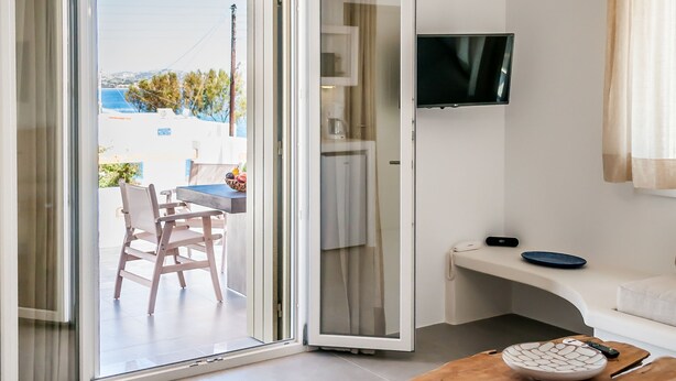 A modern living room with a sliding glass door leading to an outdoor area, a wooden coffee table, a sofa, and a flat-screen TV mounted on the wall. - Vilos Suites (Milos)
