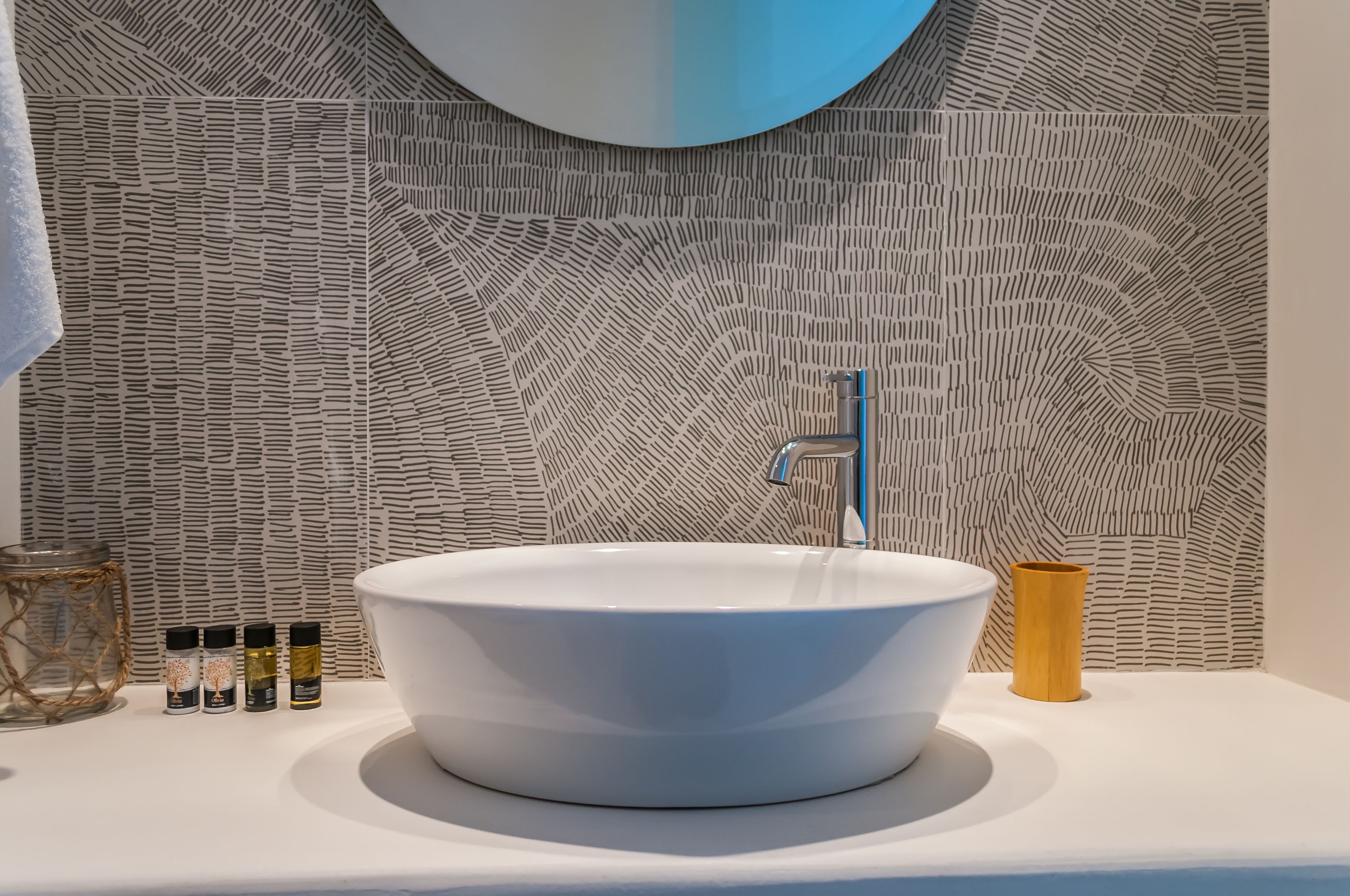 A modern bathroom with a white sink, a round mirror, and a patterned wall.