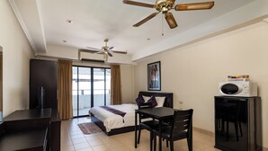 A hotel room with a bed, a desk, a chair, a ceiling fan, and a microwave. - The Classroom Hotel (Pattaya)