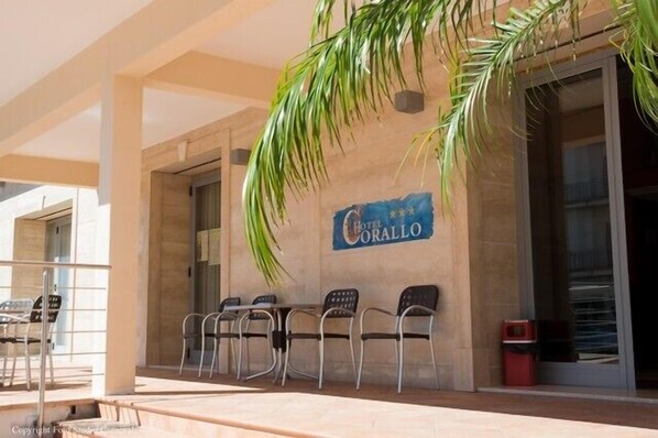 Un área de estar al aire libre con un letrero del Hotel Corallo, que presenta un diseño moderno con una palmera y una vista despejada de la entrada.