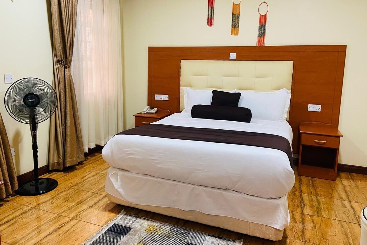 A hotel room with a large bed, a bedside table with a phone, a nightstand, a standing fan, and a wall-mounted light fixture.