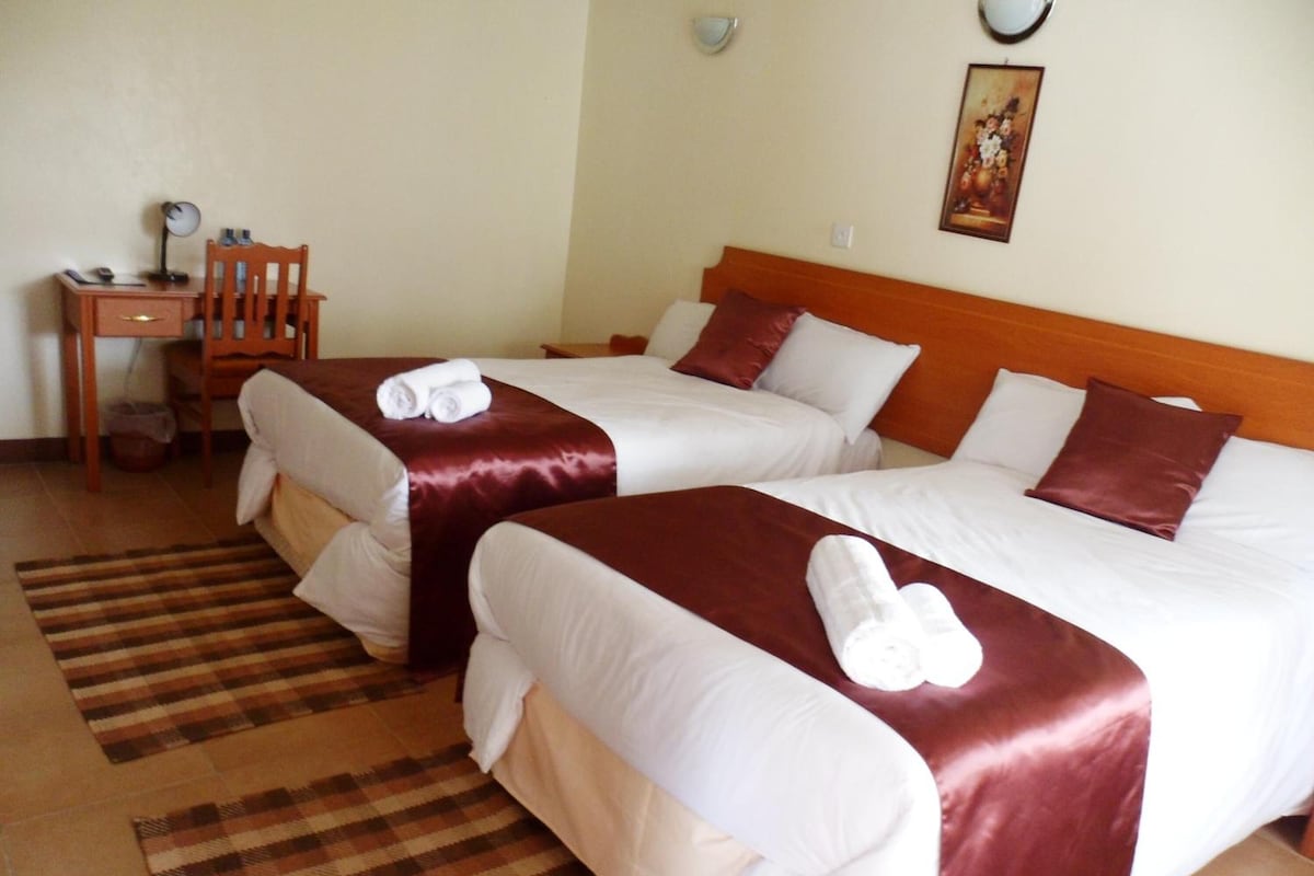 A hotel room with two beds, each with white linens and red headboards. There is a wooden chair and a small table with a lamp. A checkered rug is on the floor.
