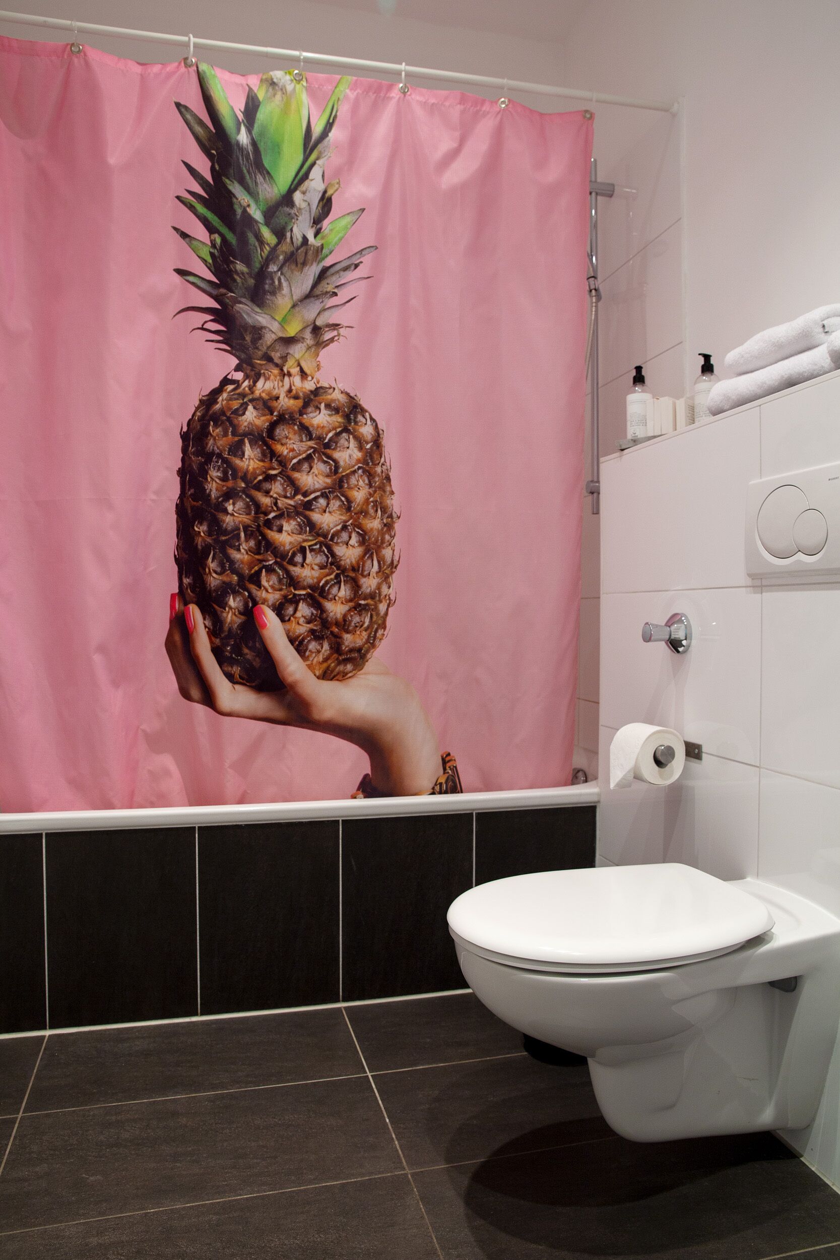 Un baño con cortina de ducha con estampado de piña, un inodoro blanco y suelo de baldosas.