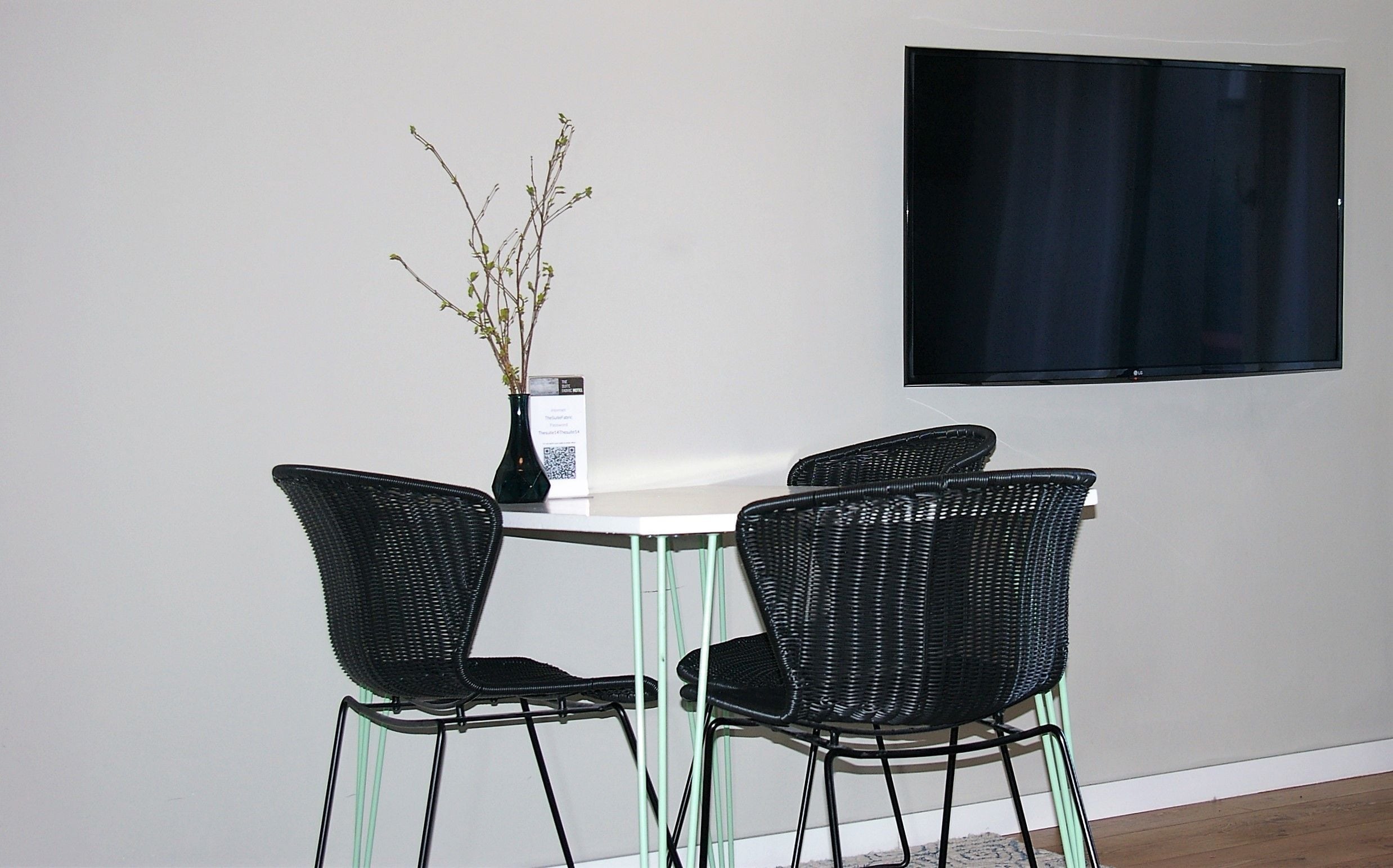 A dining area with a table and four black wicker chairs, a vase with branches, and a flat-screen TV mounted on the wall.