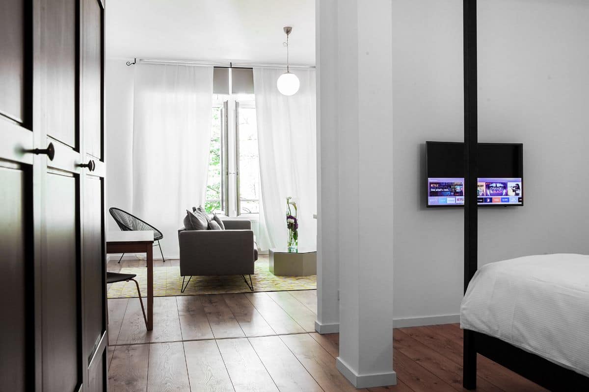 A modern bedroom with a bed, a TV mounted on the wall, a dining area with a table and chairs, and a view of the outdoors through a window.